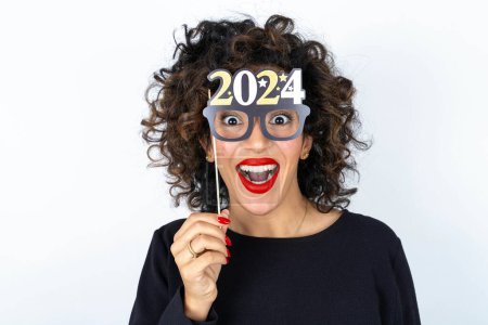 Téléchargez les photos : Jeune belle femme aux cheveux bouclés portant une robe noire sur fond de studio blanc tenant une bannière Happy New Year 2024. - en image libre de droit
