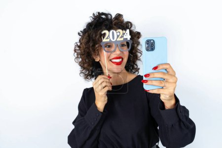 Photo for Young beautiful woman with curly hair wearing black dress, holding happy new year 2024 message and taking a picture or calling her family and friends via video call. - Royalty Free Image