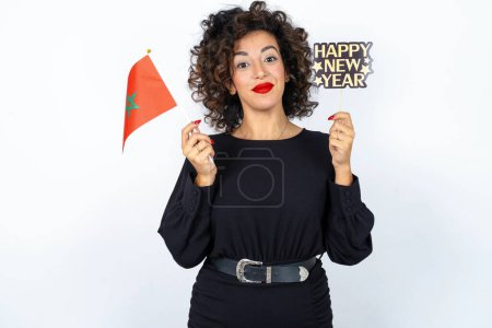 Foto de Mujer hermosa joven con el pelo rizado con un vestido negro con una bandera marroquí y una bandera feliz año nuevo sobre fondo de estudio blanco. - Imagen libre de derechos