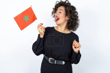 Téléchargez les photos : Jeune belle femme aux cheveux bouclés portant une robe noire et tenant un drapeau marocain se sentant confiant et heureux geste gagnant. - en image libre de droit