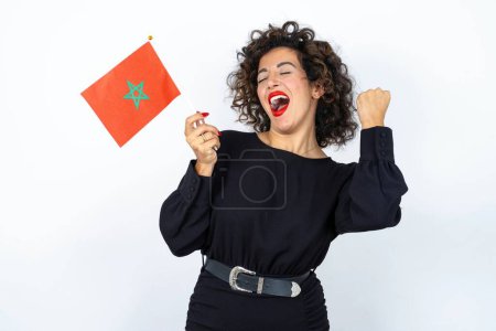 Téléchargez les photos : Jeune belle femme aux cheveux bouclés portant une robe noire et tenant un drapeau marocain se sentant confiant et heureux geste gagnant. - en image libre de droit