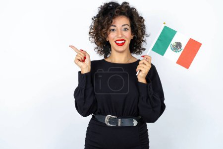 Téléchargez les photos : Jeune belle femme aux cheveux bouclés portant une robe noire et tenant un drapeau mexicain sur fond de studio blanc. - en image libre de droit
