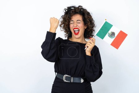 Photo for Young beautiful woman with curly hair wearing black dress and holding a Mexican flag over white studio background. - Royalty Free Image