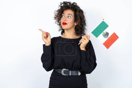 Foto de Joven hermosa mujer con el pelo rizado usando vestido negro y sosteniendo una bandera mexicana sobre fondo de estudio blanco. - Imagen libre de derechos