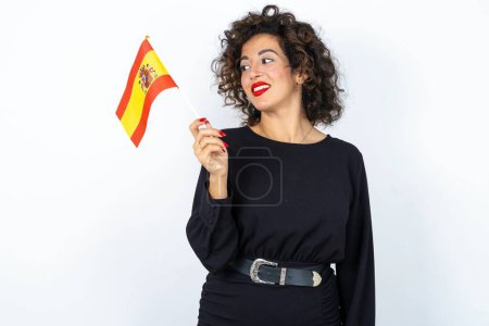 Téléchargez les photos : Jeune belle femme aux cheveux bouclés portant une robe noire et drapeau espagnol se sentant excité et confiant - en image libre de droit