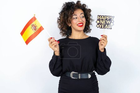 Téléchargez les photos : Jeune belle femme aux cheveux bouclés portant une robe noire, drapeau espagnol et une bannière Bonne année 2024 - en image libre de droit