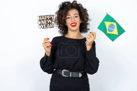 Foto de Mujer hermosa joven con el pelo rizado con vestido negro, bandera de Brasil y una bandera feliz año nuevo 2024 - Imagen libre de derechos