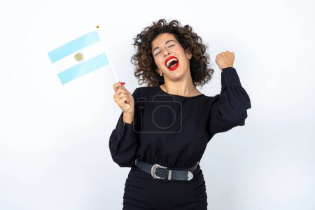 Foto de Joven hermosa mujer con el pelo rizado vistiendo vestido negro y sosteniendo una bandera argentina y sonriendo - Imagen libre de derechos