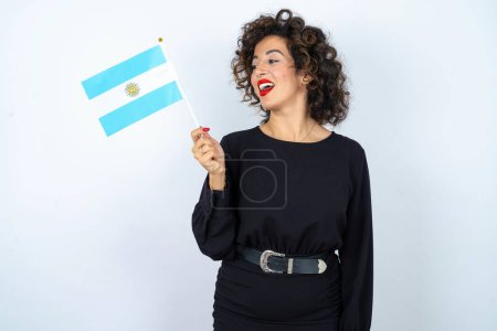 Téléchargez les photos : Jeune belle femme aux cheveux bouclés portant une robe noire et tenant un drapeau argentin et souriant - en image libre de droit