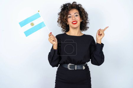 Foto de Joven hermosa mujer con el pelo rizado usando vestido negro y sosteniendo una bandera de Argentina y señalando a un lado. - Imagen libre de derechos