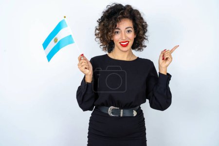 Foto de Joven hermosa mujer con el pelo rizado usando vestido negro y sosteniendo una bandera de Argentina y señalando a un lado. - Imagen libre de derechos