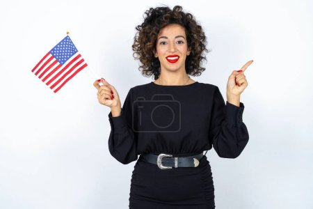 Téléchargez les photos : Jeune belle femme aux cheveux bouclés portant une robe noire et tenant et drapeau américain des États-Unis et pointant de côté sur fond de studio blanc. - en image libre de droit