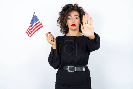 Foto de Joven hermosa mujer con el pelo rizado con vestido negro y la celebración y la bandera de Estados Unidos de América y haciendo gesto de parada sobre fondo de estudio blanco. - Imagen libre de derechos
