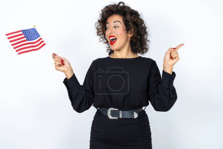 Photo for Young beautiful woman with curly hair wearing black dress and holding and American USA flag and pointing aside over white studio background. - Royalty Free Image