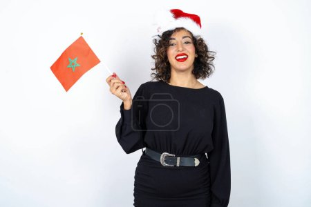 Foto de Joven hermosa mujer con el pelo rizado usando vestido negro, sombrero de Navidad y sosteniendo una bandera marroquí sonriendo y sosteniendo la mano cerca de la cara. - Imagen libre de derechos