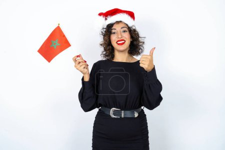 Young beautiful woman with curly hair wearing black dress, christmas hat and holding a Moroccan flag pointing up with thumb. Approval sign.