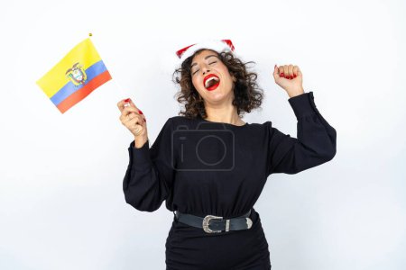Photo for Young beautiful woman with curly hair wearing black dress, christmas hat and holding a Moroccan flag smiling and holding hand near face. - Royalty Free Image