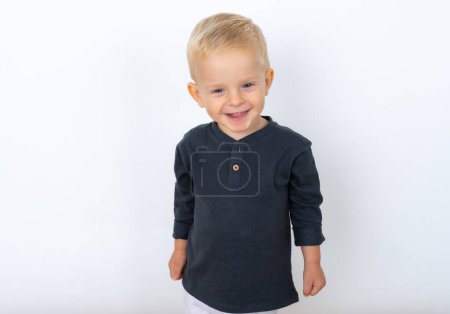 Téléchargez les photos : Plan studio de garçon blond heureux enfant portant t-shirt noir et pantalon blanc posant sur fond de studio blanc - en image libre de droit