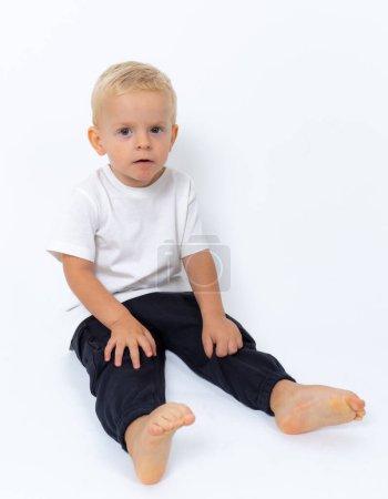 Photo for Blond kid boy wearing white t-shirt and black pants sitting on the floor and posing over white studio background - Royalty Free Image