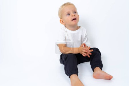 Photo for Blond kid boy wearing white t-shirt and black pants sitting on the floor and posing over white studio background - Royalty Free Image