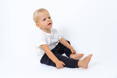Téléchargez les photos : Garçon blond enfant portant un t-shirt blanc et un pantalon noir assis sur le sol et posant sur fond de studio blanc - en image libre de droit