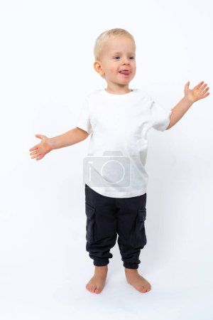 Foto de Retrato del hermoso niño rubio con camiseta blanca y pantalones negros posando sobre fondo blanco del estudio - Imagen libre de derechos