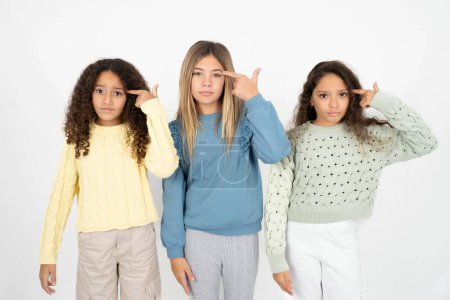 Photo for Unhappy three teenager girls makes suicide gesture and imitates gun with hand, curves lips keeps two fingers on temple, shoots, being tired of everything, - Royalty Free Image