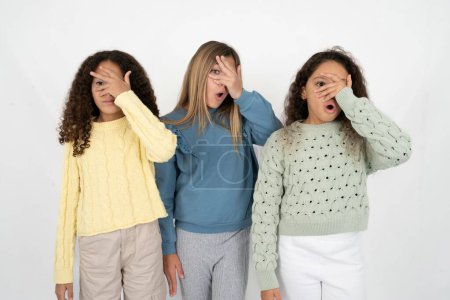 Foto de Tres chicas adolescentes asomándose en shock cubriendo la cara y los ojos con la mano, mirando a través de los dedos con expresión avergonzada. - Imagen libre de derechos