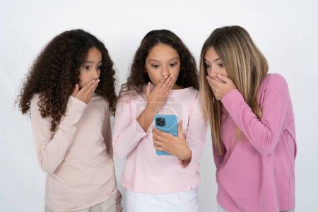 Foto de Tres adolescentes que se sorprenden profundamente, mira fijamente a la pantalla del teléfono inteligente, lee noticias impactantes en el sitio web, Omg, su horrible! - Imagen libre de derechos