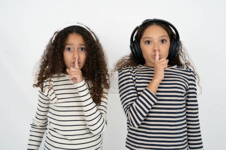 Photo for Multiracial group of girl friends over white studio background making hush gesture with finger on her lips wearing  wireless headphones. Be quiet. - Royalty Free Image