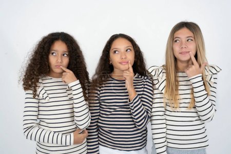 Foto de Encantador dreamy tres adolescente niñas mantiene dedo cerca de labios looks a un lado copia espacio. - Imagen libre de derechos