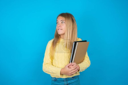 Photo for Amazed beautiful kid girl wearing yellow sweater over blue background biting lip and looking tricky to empty space. - Royalty Free Image