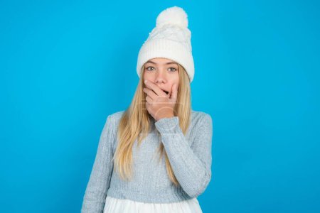 Photo for Emotional beautiful kid girl wearing white knitted hat and blue sweater gasps from astonishment, covers opened mouth with palm, looks shocked at camera. - Royalty Free Image