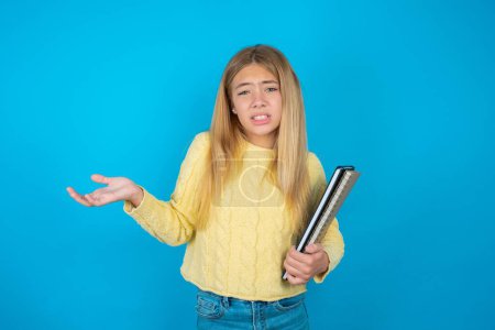 Photo for Clueless beautiful kid girl wearing yellow sweater over blue background shrugs shoulders with hesitation, faces doubtful situation, spreads palms, Hard decision - Royalty Free Image