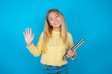 Foto de Hermosa niña vistiendo suéter amarillo sobre fondo azul saludando feliz y sonriente gesto de bienvenida. - Imagen libre de derechos