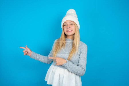 Foto de Hermosa niña con sombrero de punto blanco y suéter azul que indica el dedo espacio vacío que muestra los mejores precios bajos, mirando a la cámara - Imagen libre de derechos