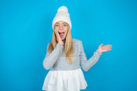 Photo for Crazy beautiful kid girl wearing white knitted hat and blue sweater advising discount prices hold open palm new product - Royalty Free Image