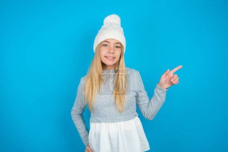 Foto de Hermosa niña con sombrero de punto blanco y suéter azul mirando a la cámara que indica las ventas de espacio vacío dedo - Imagen libre de derechos