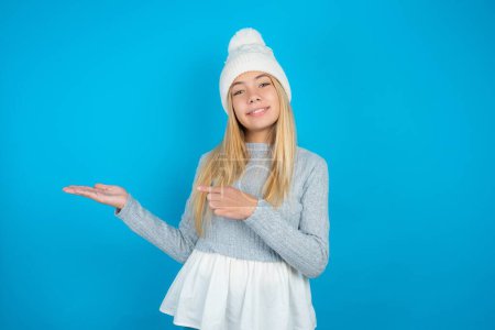 Foto de Hermosa niña con sombrero de punto blanco y suéter azul apuntando y sosteniendo la mano mostrando anuncios - Imagen libre de derechos