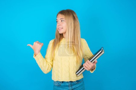 Charming beautiful kid girl wearing yellow sweater over blue background looking at copy space having advertisements