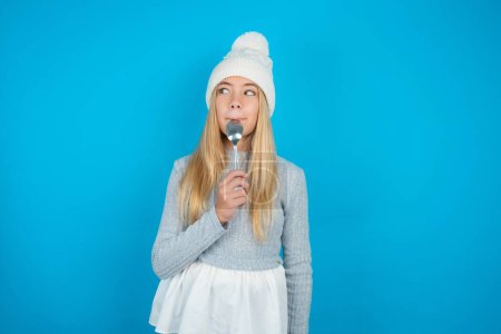 Photo for Very hungry beautiful kid girl wearing white knitted hat and blue sweater holding spoon into mouth dream of tasty meal - Royalty Free Image