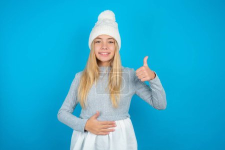 Foto de Hermosa niña con sombrero de punto blanco y suéter azul feliz sonrisa positiva manos en el vientre mostrar pulgar hacia arriba bien saludable - Imagen libre de derechos