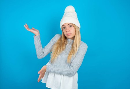 Téléchargez les photos : Belle gamine portant chapeau tricoté blanc et pull bleu pointant de côté avec les deux mains montrant quelque chose d'étrange et disant : Je ne sais pas ce que c'est. Concept de publicité. - en image libre de droit