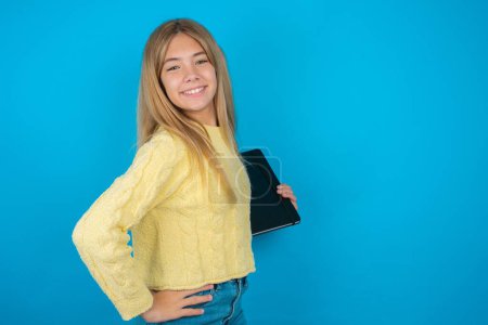 beautiful kid girl wearing yellow sweater over blue background feeling happy and cheerful, smiling and welcoming you, inviting you in with a friendly gesture