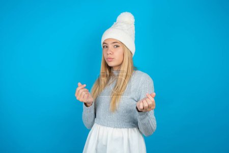 Foto de Hermosa niña vistiendo sombrero de punto blanco y suéter azul haciendo gesto de dinero con las manos, pidiendo pago de sueldo, negocio millonario - Imagen libre de derechos
