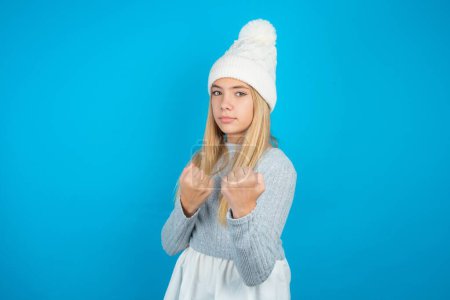 Photo for Beautiful kid girl wearing white knitted hat and blue sweater Ready to fight with fist defense gesture, angry and upset face, afraid of problem. - Royalty Free Image