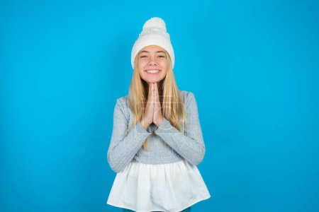 Téléchargez les photos : Belle enfant fille portant chapeau tricoté blanc et pull bleu priant avec les mains ensemble demandant pardon souriant confiant. - en image libre de droit
