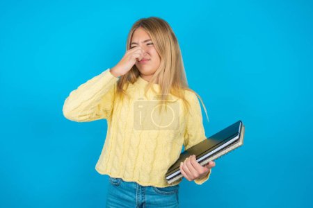 Photo for Beautiful kid girl wearing yellow sweater over blue background smelling something stinky and disgusting, intolerable smell, holding breath with fingers on nose. Bad smell - Royalty Free Image