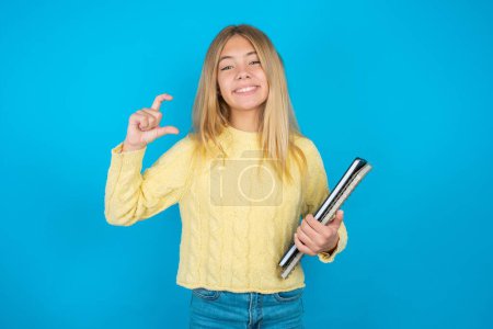 Téléchargez les photos : Belle enfant fille portant un pull jaune sur fond bleu geste souriant et confiant avec la main faisant signe de petite taille avec les doigts à la recherche et la caméra. Concept de mesure - en image libre de droit