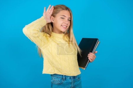 Foto de Hermosa niña vistiendo suéter amarillo sobre fondo azul tratando de escuchar ambas manos en el gesto de la oreja, curioso por chismes. Problema de audición, sordos - Imagen libre de derechos